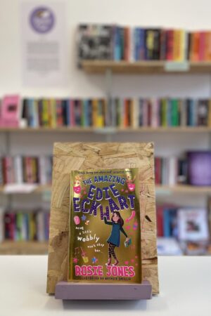 Copi o 'The Amazing Edie Eckhart' yn sefyll ar stondin llyfrau, tu blaen silffoedd o lyfrau yn y cefndir.