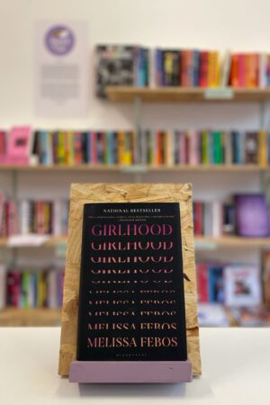 Copi o 'Girlhood' yn sefyll ar stondin llyfrau, tu blaen silffoedd o lyfrau yn y cefndir.