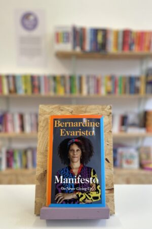 Copi o 'Manifesto' yn sefyll ar stondin llyfrau, tu blaen silffoedd o lyfrau yn y cefndir.