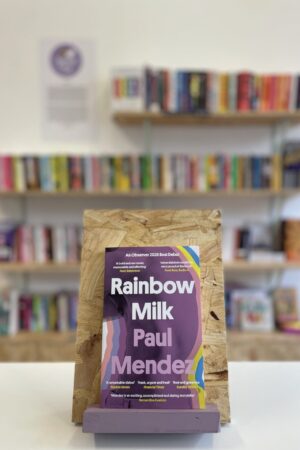 Cymraeg: Copi o 'Rainbow Milk' yn sefyll ar stondin llyfrau, tu blaen silffoedd o lyfrau yn y cefndir. | English: A copy of 'Rainbow Milk' sits on a stand in front of multiple shelves of other books.