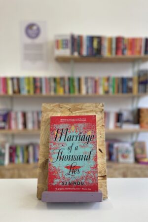 Copi o 'Marriage of a Thousand Lies' yn sefyll ar stondin llyfrau, tu blaen silffoedd o lyfrau yn y cefndir.