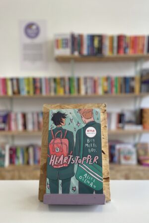 Copi o 'Heartstopper vol. 1' yn sefyll ar stondin llyfrau, tu blaen silffoedd o lyfrau yn y cefndir.