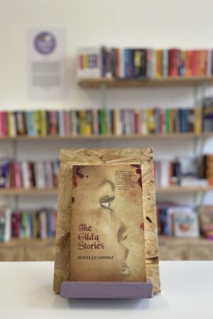 Cymraeg: Copi o 'The Gilda Stories' yn sefyll ar stondin llyfrau, tu blaen silffoedd o lyfrau yn y cefndir. | English: A copy of 'The Gilda Stories' sits on a stand in front of multiple shelves of other books.