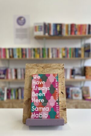 Copi o 'We Have Always Been Here' yn sefyll ar stondin llyfrau, tu blaen silffoedd o lyfrau yn y cefndir.