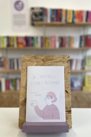 Cymraeg: Copi o 'A Potted Trans History' yn sefyll ar stondin llyfrau, tu blaen silffoedd o lyfrau yn y cefndir. English: A copy of 'A Potted Trans History' sits on a stand in front of multiple shelves of other books.