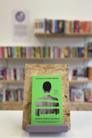 Copi o 'Her Body & Other Parties' yn sefyll ar stondin llyfrau, tu blaen silffoedd o lyfrau yn y cefndir.