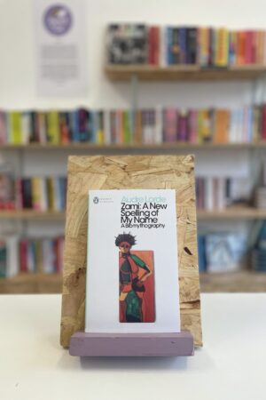 Copi o 'Zami' yn sefyll ar stondin llyfrau, tu blaen silffoedd o lyfrau yn y cefndir.