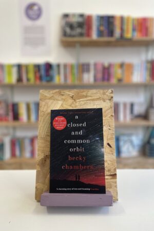 Copi o 'A Closed and Common Orbit' yn sefyll ar stondin llyfrau, tu blaen silffoedd o lyfrau yn y cefndir.