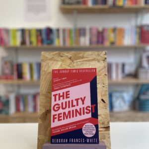Cymraeg: Copi o 'The Guilty Feminist' yn sefyll ar stondin llyfrau, tu blaen silffoedd o lyfrau yn y cefndir. | English: A copy of 'The Guilty Feminist' sits on a stand in front of multiple shelves of other books.