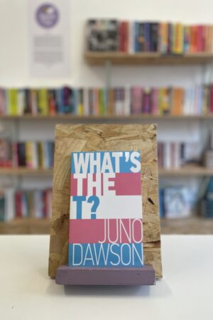 Copi o 'What's the T?' yn sefyll ar stondin llyfrau, tu blaen silffoedd o lyfrau yn y cefndir.