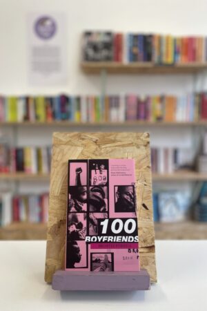Cymraeg: Copi o '100 Boyfriends' yn sefyll ar stondin llyfrau, tu blaen silffoedd o lyfrau yn y cefndir. | English: A copy of '100 Boyfriends' sits on a stand in front of multiple shelves of other books.