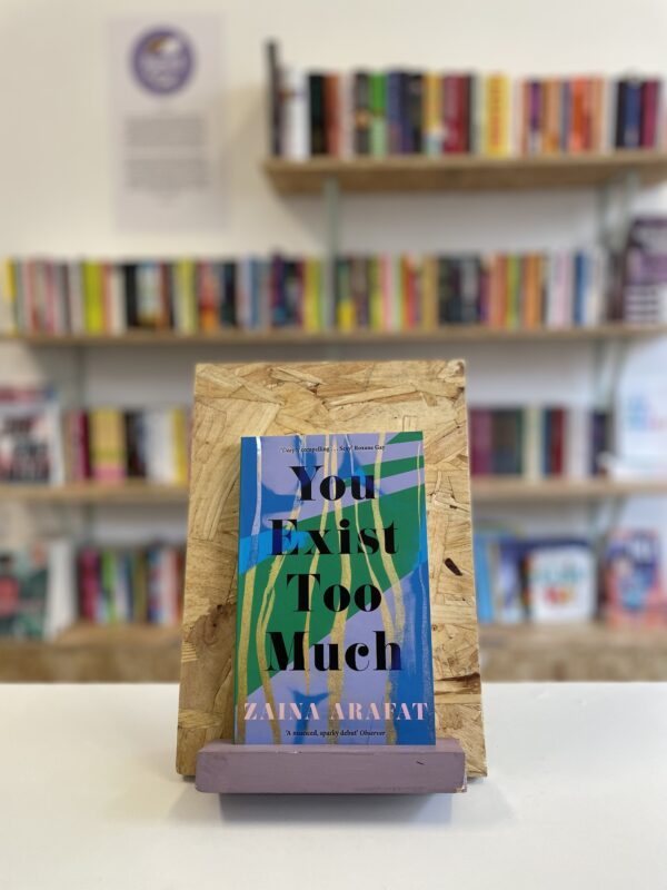 Cymraeg: Copi o 'You Exist Too Much' yn sefyll ar stondin llyfrau, tu blaen silffoedd o lyfrau yn y cefndir. | English: A copy of 'You Exist Too Much' sits on a stand in front of multiple shelves of other books.