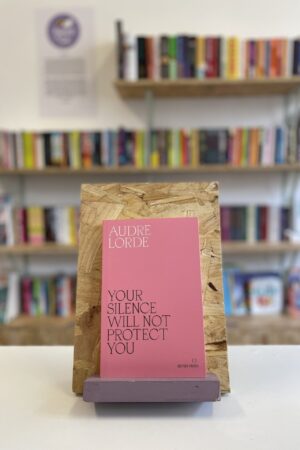 Copi o 'Your Silence Will Not Protect You' yn sefyll ar stondin llyfrau, tu blaen silffoedd o lyfrau yn y cefndir.