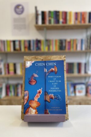 Copi o 'When I Grow Up I Want To Be A List of Further Possibilities' yn sefyll ar stondin llyfrau, tu blaen silffoedd o lyfrau yn y cefndir.