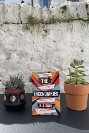 Rhyngddo dau blanhigyn mewn potiau gwahanol ar fwrdd du, mae copi o'r llyfr yn sefyll, gydag wal wen yn y cefndir.