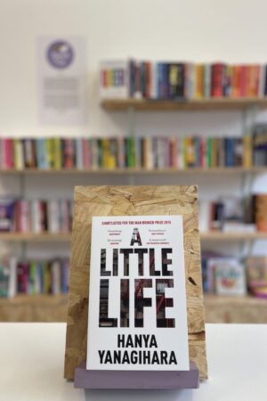 Copi o 'A Little Life' yn sefyll ar stondin llyfrau, tu blaen silffoedd o lyfrau yn y cefndir.