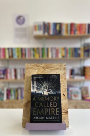 Copi o 'A Memory Called Empire' yn sefyll ar stondin llyfrau, tu blaen silffoedd o lyfrau yn y cefndir.