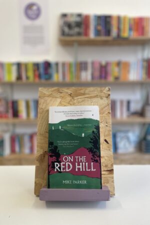 Copi o 'On the Red Hill' yn sefyll ar stondin llyfrau, tu blaen silffoedd o lyfrau yn y cefndir.