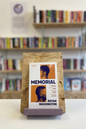 Cymraeg: Copi o 'Memorial' yn sefyll ar stondin llyfrau, tu blaen silffoedd o lyfrau yn y cefndir. | English: A copy of 'Memorial' sits on a stand in front of multiple shelves of other books.