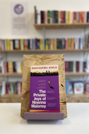 Copi o 'The Private Joys of Nnenna Maloney' yn sefyll ar stondin llyfrau, tu blaen silffoedd o lyfrau yn y cefndir.