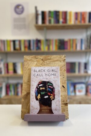 Copi o 'Black Girl, Call Home' yn sefyll ar stondin llyfrau, tu blaen silffoedd o lyfrau yn y cefndir.