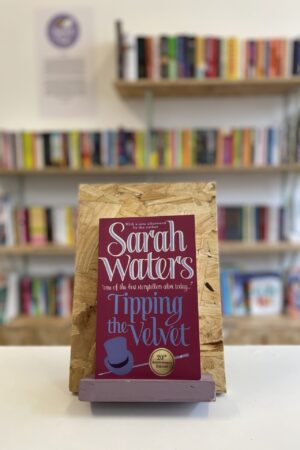 Copi o 'Tipping the Velvet' yn sefyll ar stondin llyfrau, tu blaen silffoedd o lyfrau yn y cefndir.
