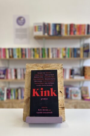 Copi o 'Kink' yn sefyll ar stondin llyfrau, tu blaen silffoedd o lyfrau yn y cefndir.