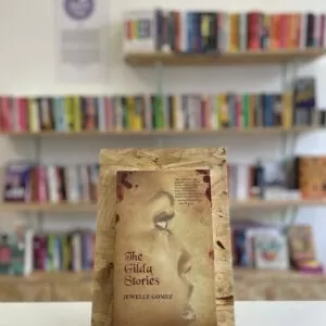 Cymraeg: Copi o 'The Gilda Stories' yn sefyll ar stondin llyfrau, tu blaen silffoedd o lyfrau yn y cefndir. | English: A copy of 'The Gilda Stories' sits on a stand in front of multiple shelves of other books.