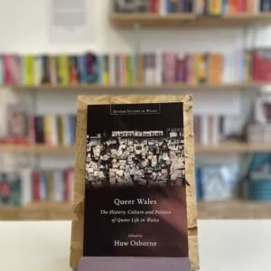 Copi o 'Queer Wales' yn sefyll ar stondin llyfrau, tu blaen silffoedd o lyfrau yn y cefndir.