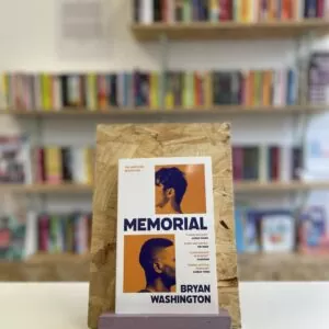 Cymraeg: Copi o 'Memorial' yn sefyll ar stondin llyfrau, tu blaen silffoedd o lyfrau yn y cefndir. | English: A copy of 'Memorial' sits on a stand in front of multiple shelves of other books.