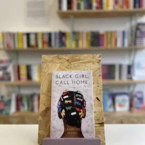 Copi o 'Black Girl, Call Home' yn sefyll ar stondin llyfrau, tu blaen silffoedd o lyfrau yn y cefndir.
